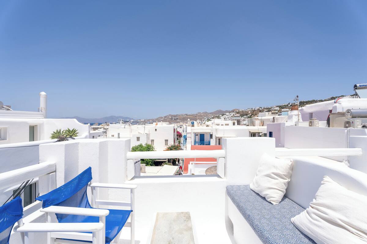 Una terraza en la azotea con mobiliario blanco y azul, vista a un pueblo con edificios blancos y un cielo azul despejado.