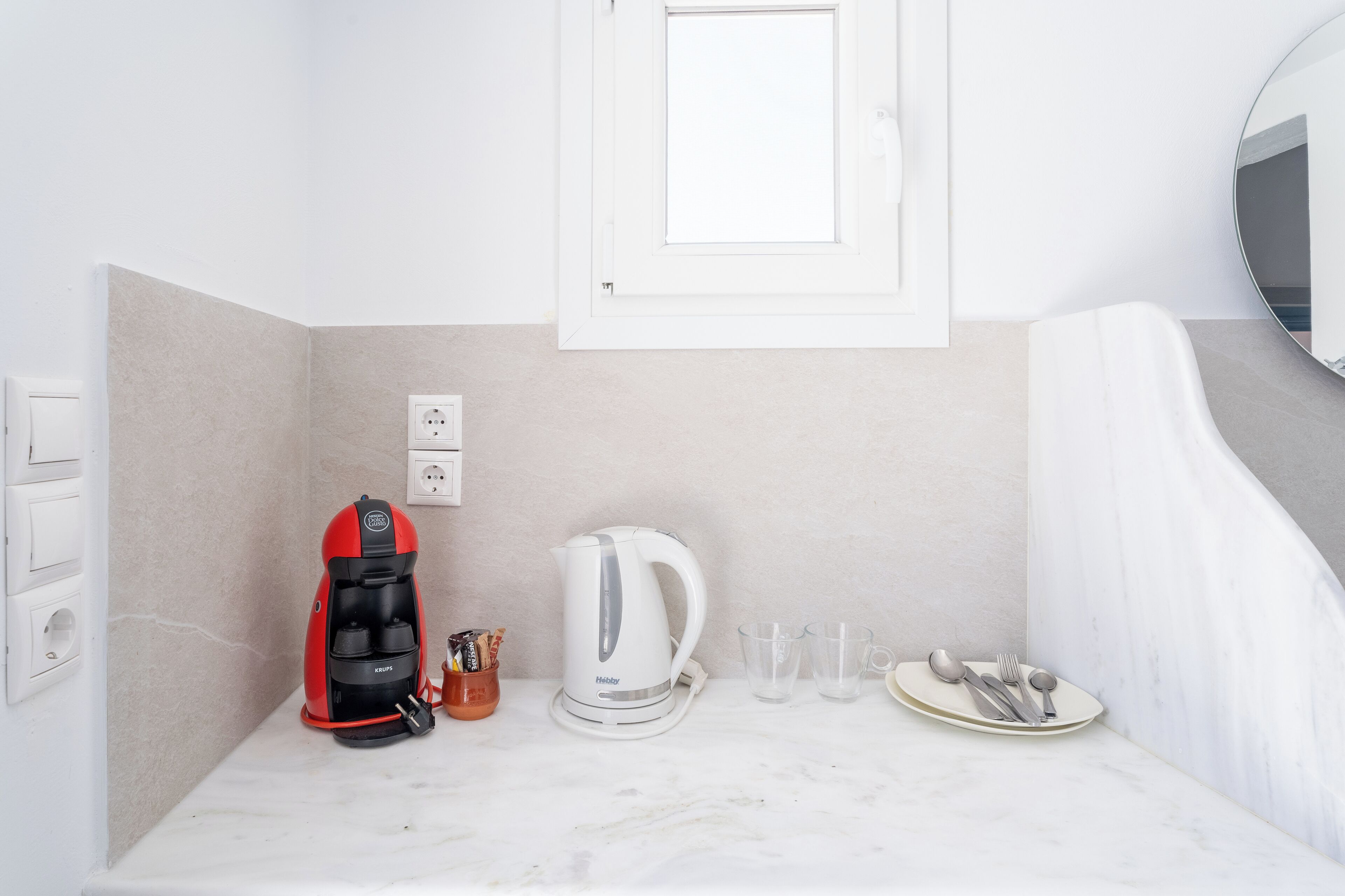 Un baño moderno con encimera de mármol blanco, cafetera roja y negra, hervidor blanco y un plato con cubiertos.