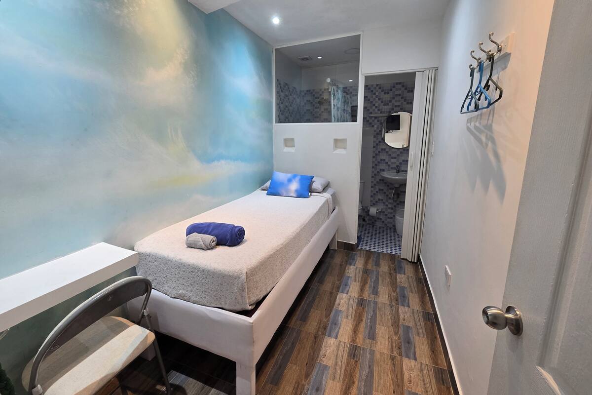 A small, modern room with a bed, a chair, a bathroom with a mirror and sink, and a wall with a blue pattern.