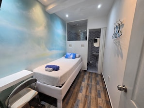 Une petite chambre moderne, équipée d’un lit, d’une chaise, d’une salle de bain avec un miroir et un lavabo, et d’un mur orné d’un motif bleu.