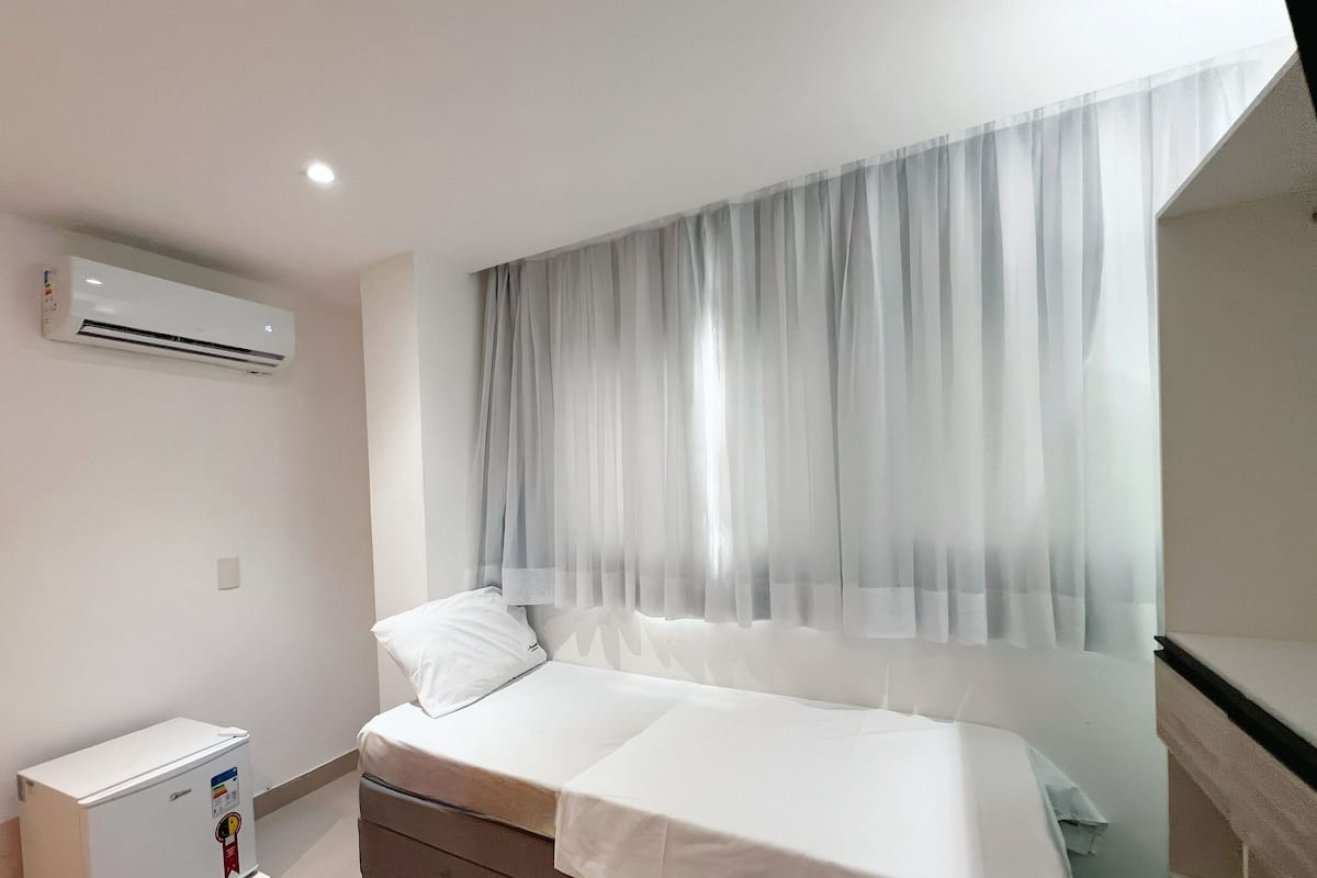 A bedroom with a bed, a window with sheer curtains, an air conditioning unit, and a small bedside table.