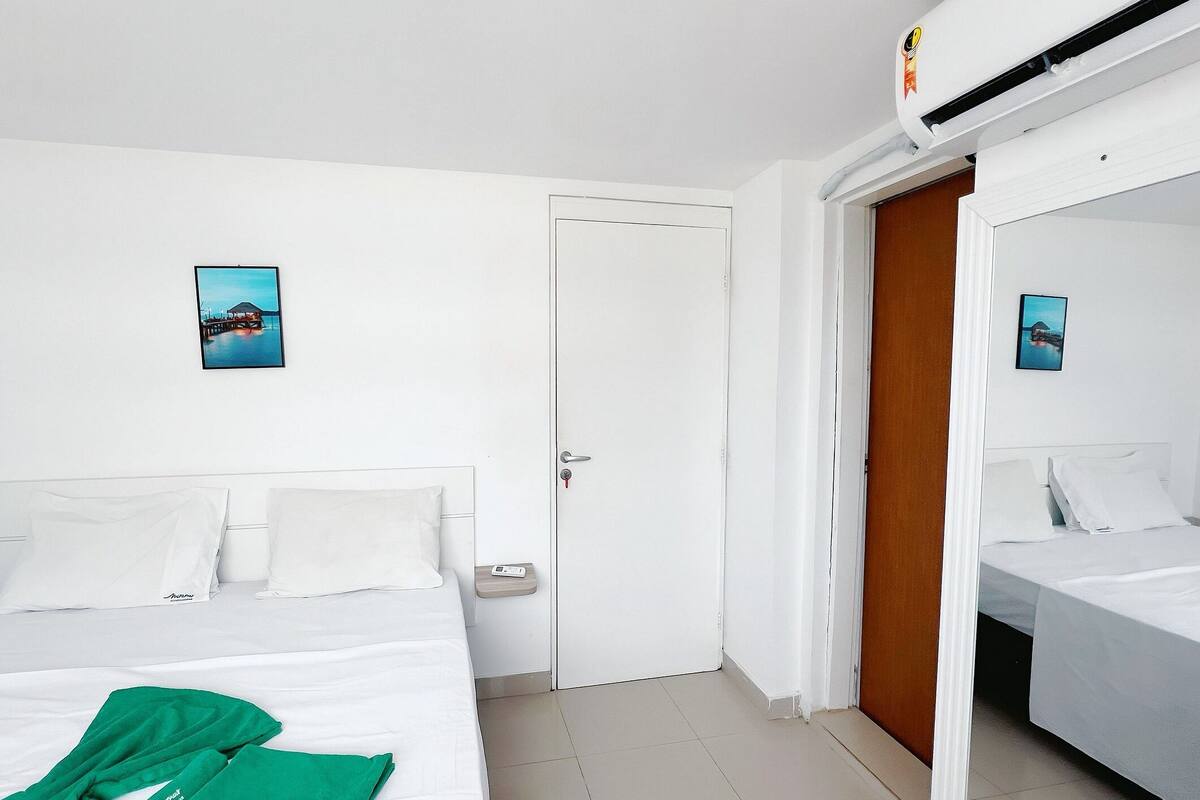 A hotel room with a bed, a nightstand, a wall-mounted air conditioner, and a door with a mirror.