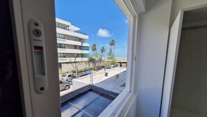 A window view of a modern building with a clear sky, palm trees, and parked cars. - Miramar Na Praia (João Pessoa)