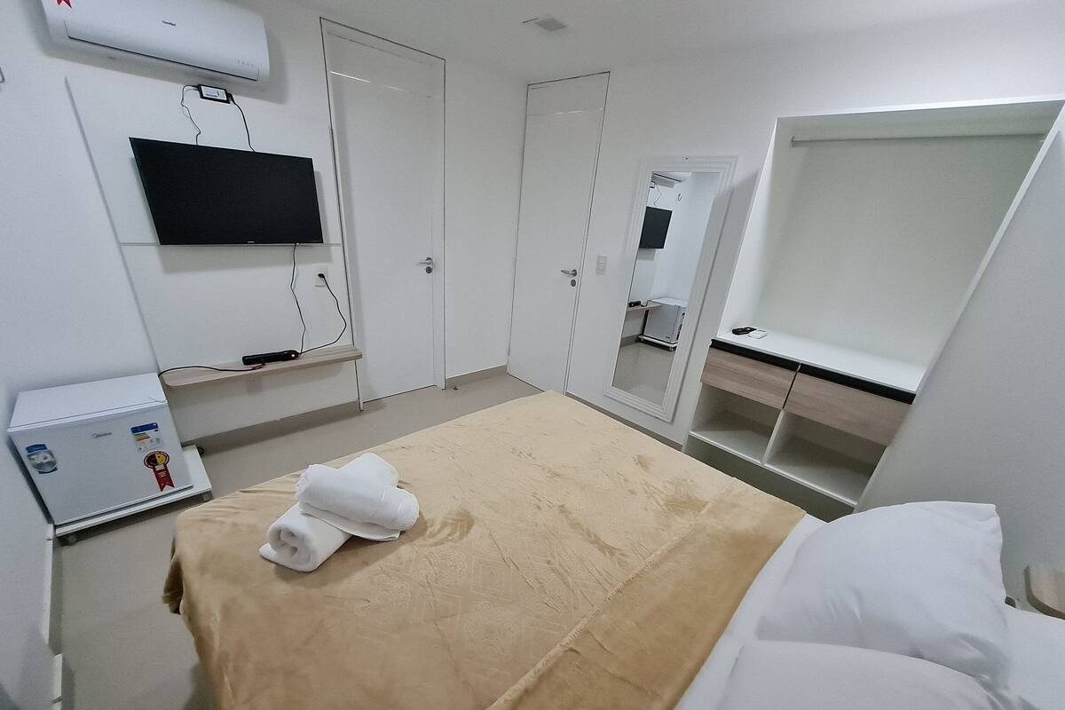 A hotel room with a bed, a television mounted on the wall, a minibar, and a door leading to another room.