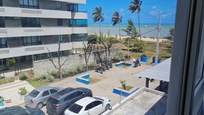 A modern building with multiple floors, a parking area with several cars, and a view of the beach with palm trees and the ocean. - Miramar Na Praia (João Pessoa)