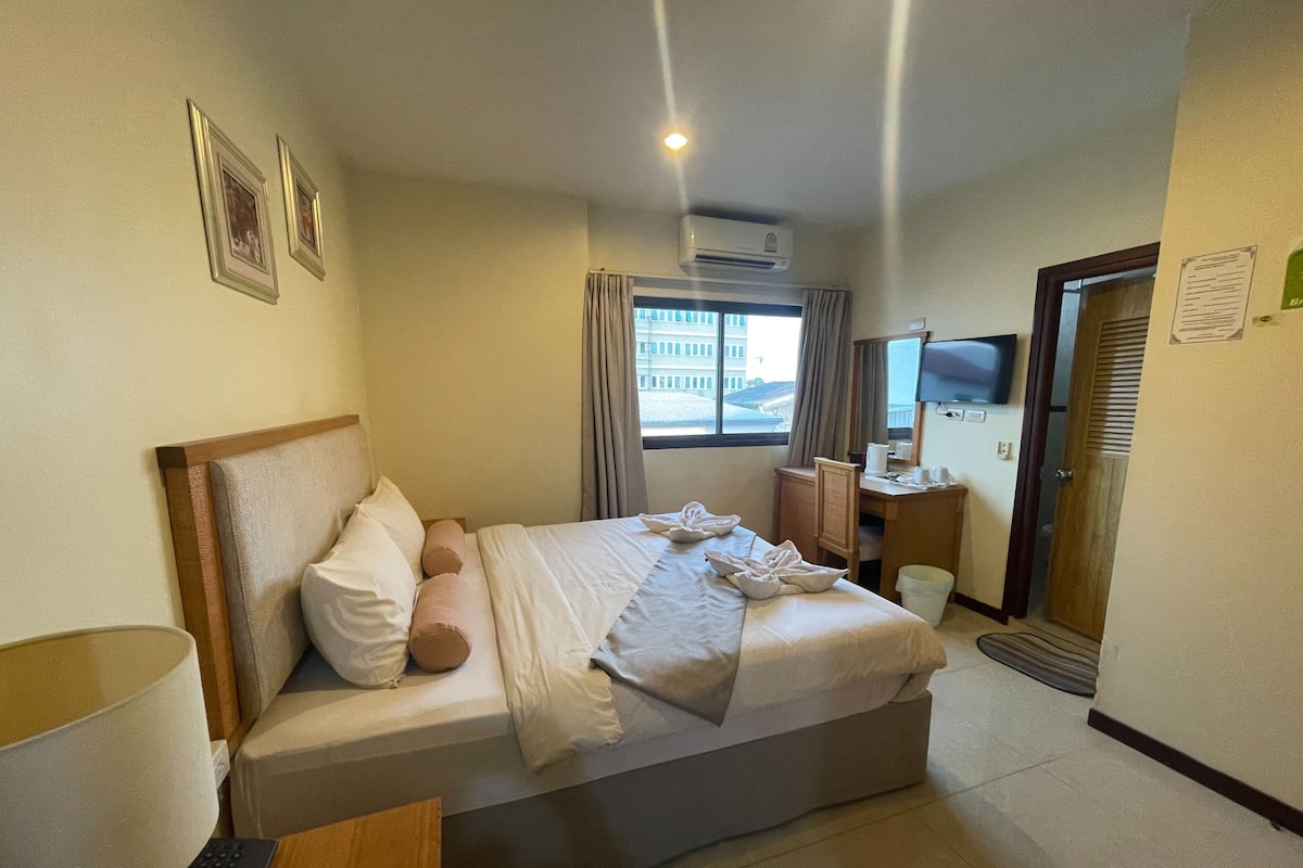 A hotel room with a bed, bedside table, lamp, air conditioning unit, window with curtains, and a television.