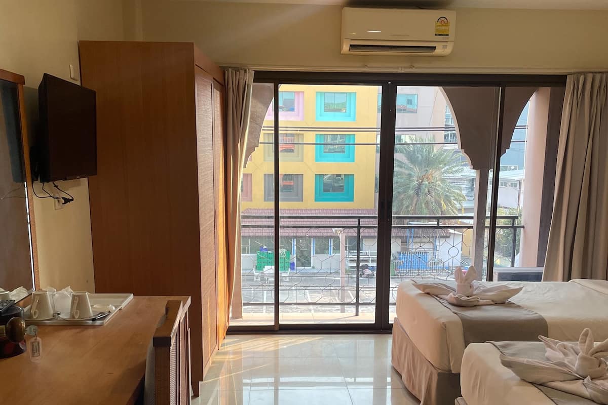A hotel room with a wooden desk, two beds, a TV, and a balcony with a view of colorful buildings.