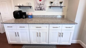 A kitchen with white cabinets, a granite countertop, and a coffee maker. There are two shelves above the countertop holding glassware and a decorative piece. - Adorable 1 BR on the mount (Pittsburgh)