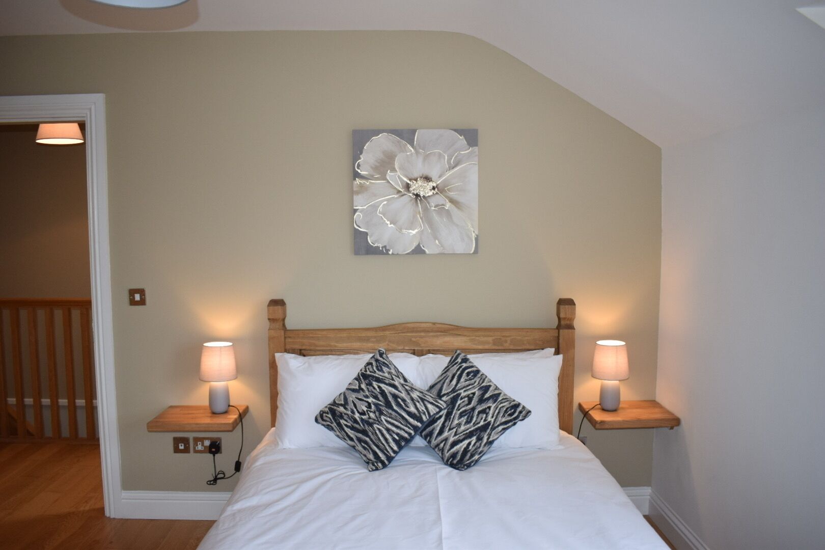 A bedroom with a wooden headboard, two bedside lamps, a white bedspread, and a large floral artwork on the wall.