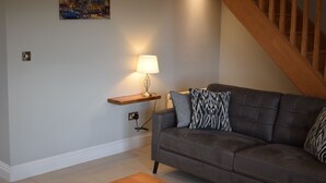 A living room with a dark brown sofa, a wooden shelf with a lamp, and a painting of a cityscape.