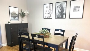 Un comedor con una mesa de madera y cuatro sillas, un florero con flores blancas y tres cuadros en la pared.