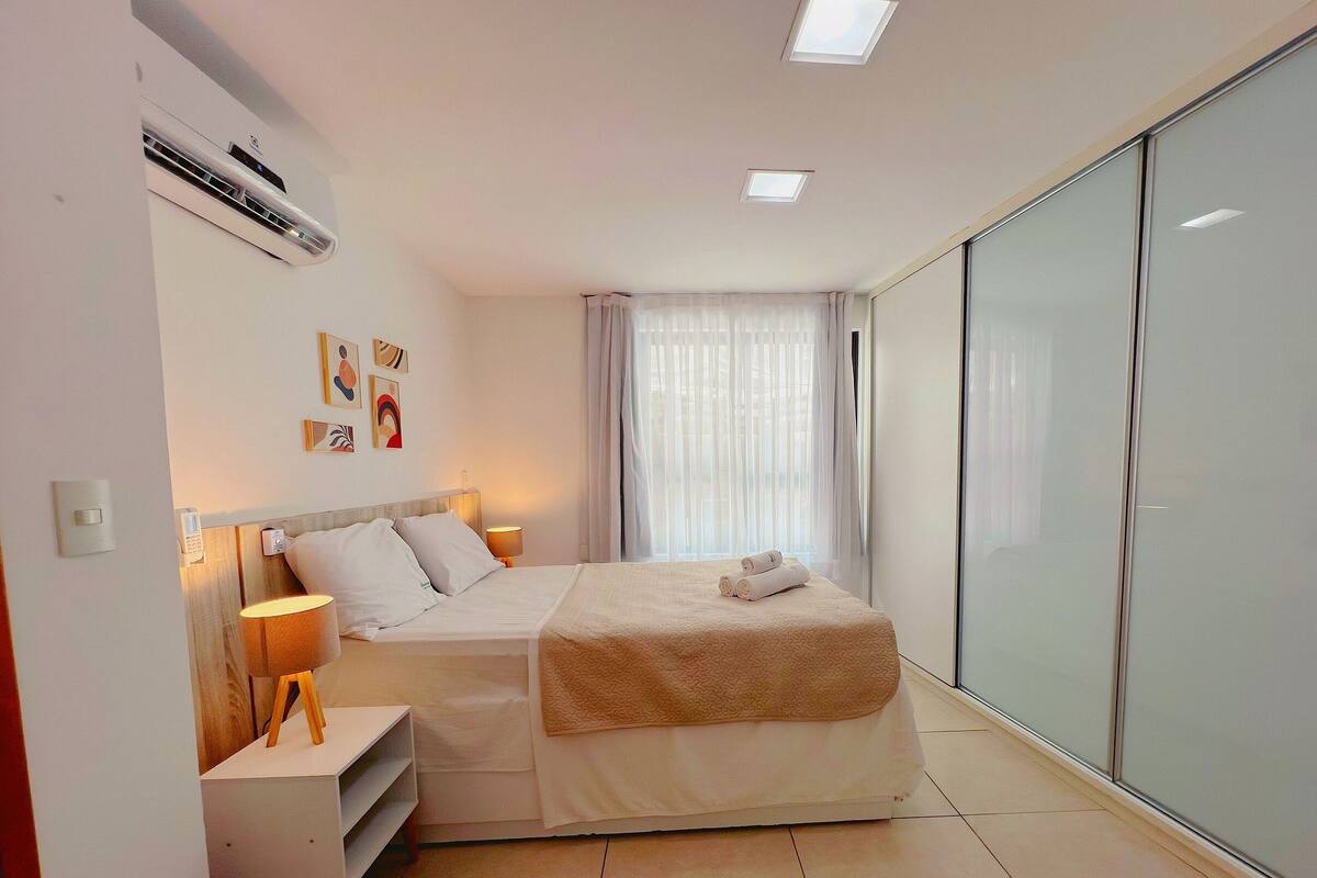 A modern bedroom with a bed, bedside tables, a lamp, artwork on the wall, and a sliding glass door closet.