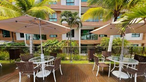 A rooftop terrace with outdoor seating, palm trees, and a modern building in the background. - Miramar Luxor Paulo Miranda - C. Branco (João Pessoa)