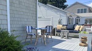 Una terraza de madera con mobiliario exterior, incluyendo una mesa y sillas, y un pequeño jardín con plantas y flores.