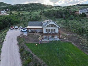 A large house with a covered porch, a lawn, and a dirt road with boats and vehicles. - Kozy Retreat @ Bear Lake - 6 KING Beds, Bunk Room, Lake View, Deck w/ Grill (Garden City)