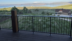 Una terraza de madera con barandal metálico que da a un paisaje cubierto de hierba, con una casa y una masa de agua al fondo.