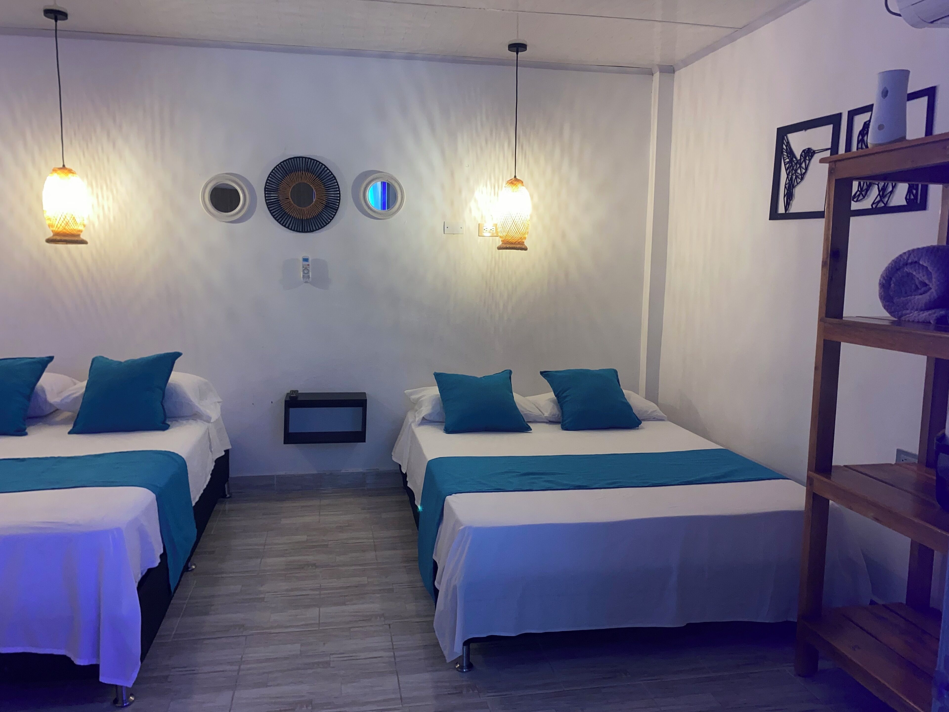 Habitación de hotel con dos camas, cada una con ropa de cama y almohadas color turquesa, una mesita de noche pequeña y un estante en la pared con una toalla enrollada.
