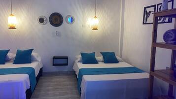 Habitación de hotel con dos camas, cada una con ropa de cama y almohadas color turquesa, una mesita de noche pequeña y un estante en la pared con una toalla enrollada.