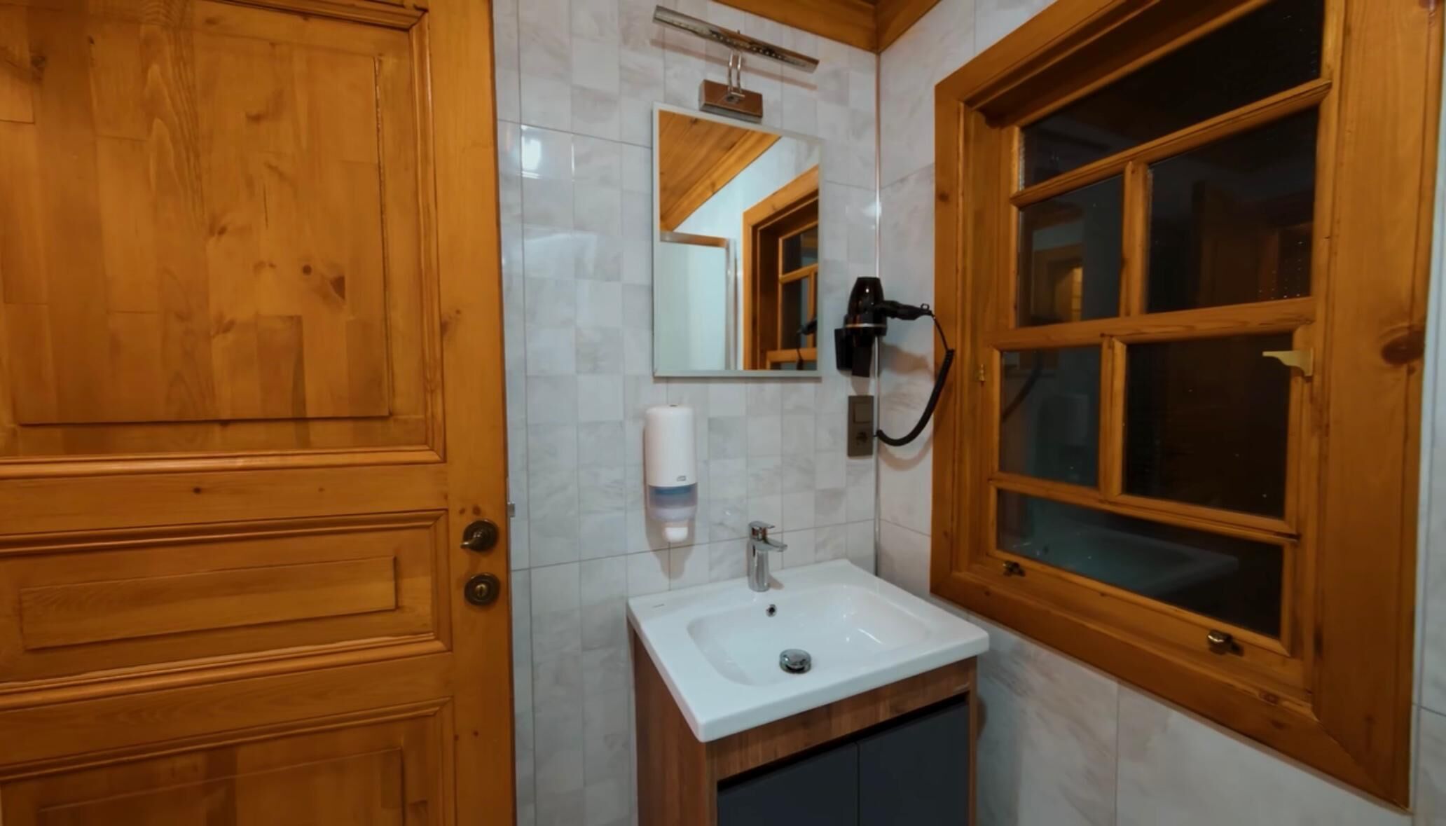 Eine Holztür, ein weißes Waschbecken mit Spiegel darüber und ein Fenster mit Holzrahmen.