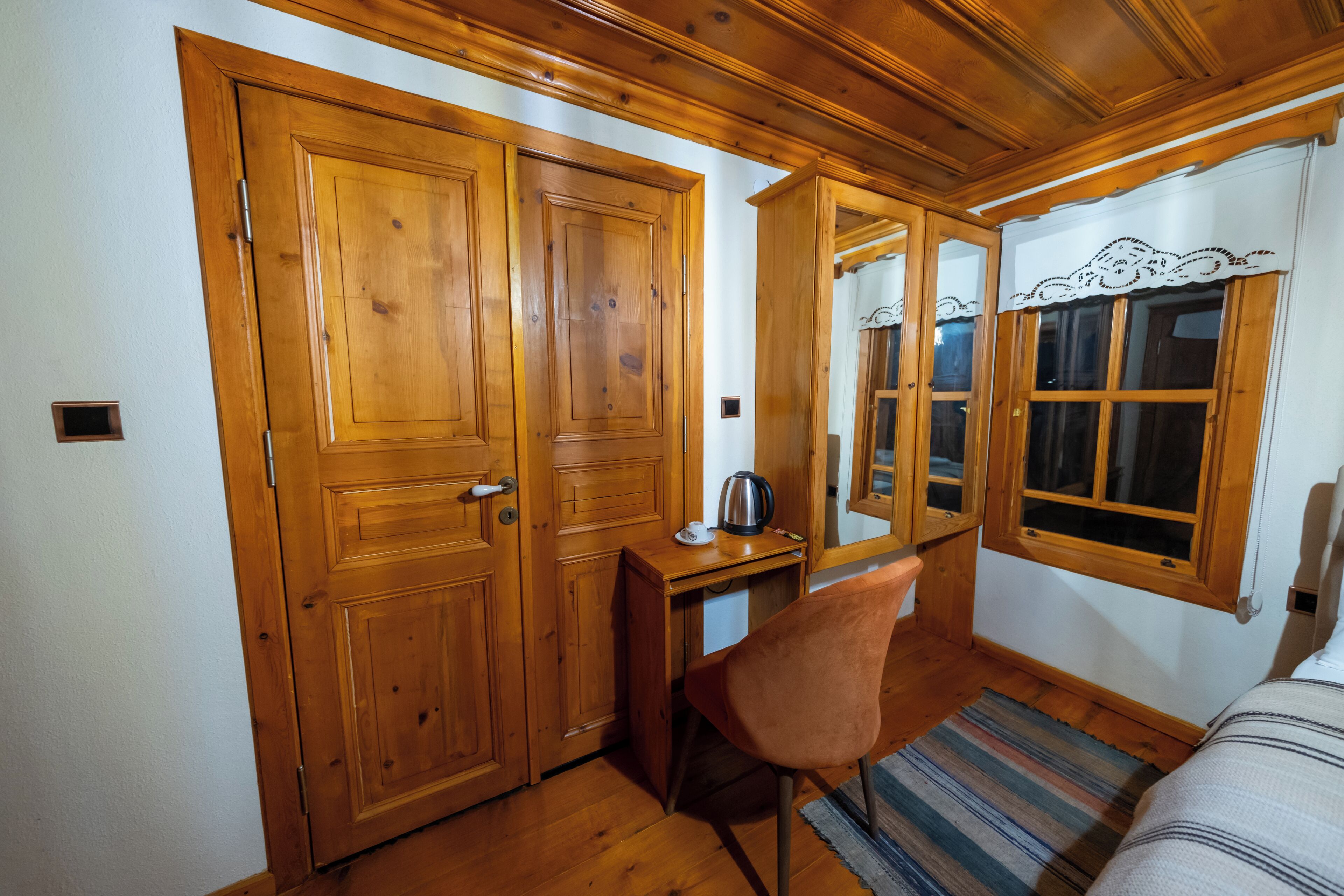 Una habitación con puertas de madera, un escritorio con una silla, una cama y una ventana con cortinas.