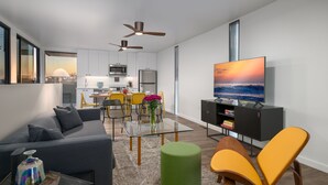 A modern living room with a sofa, a glass coffee table, a green ottoman, and a TV on a black stand. There is a kitchen in the background with a dining area. - 4 Bed Penthouse w/City View Deck and Firepit. Near Zoo (San Diego)