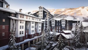 Ein mehrstöckiges Hotel mit modernem architektonischen Design, das dunkle und helle Fassaden kombiniert und von schneebedeckten Bäumen und einer Bergkulisse umgeben ist.