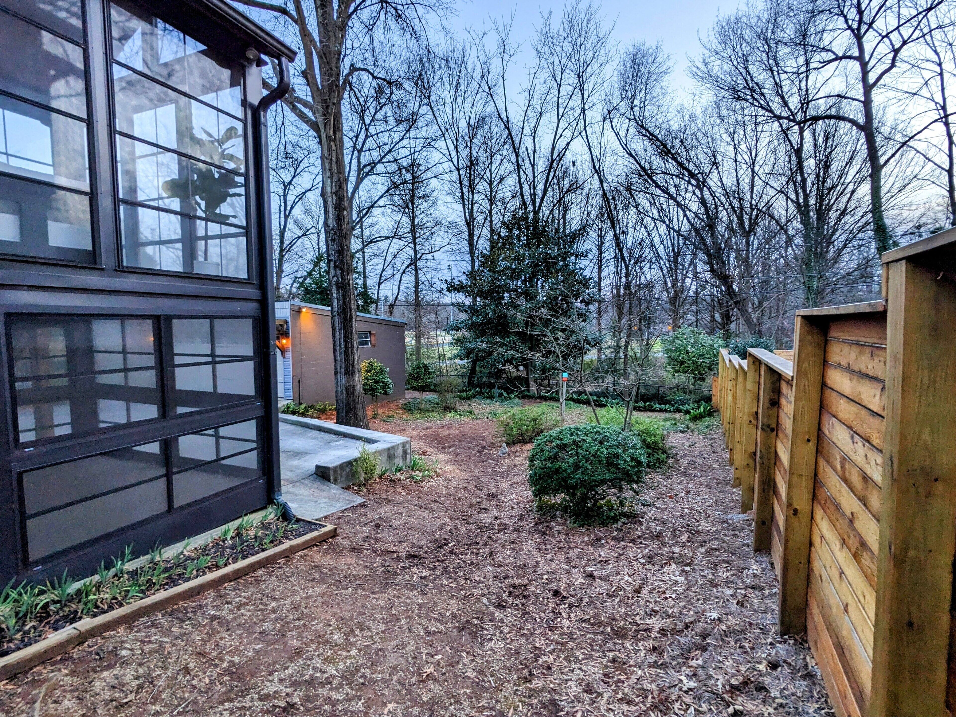 A modern house with a large glass window, a wooden fence, and a well-maintained garden with a small tree and shrubs.