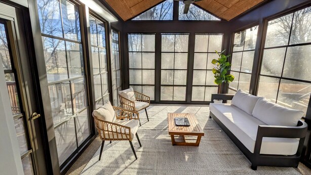 A sunroom with large windows, a ceiling fan, wicker chairs, a white sofa, and a wooden coffee table.