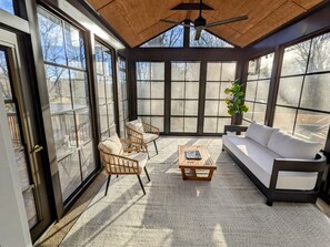 Une véranda avec de grandes fenêtres, un ventilateur de plafond, des fauteuils en osier, un canapé blanc et une table basse en bois.