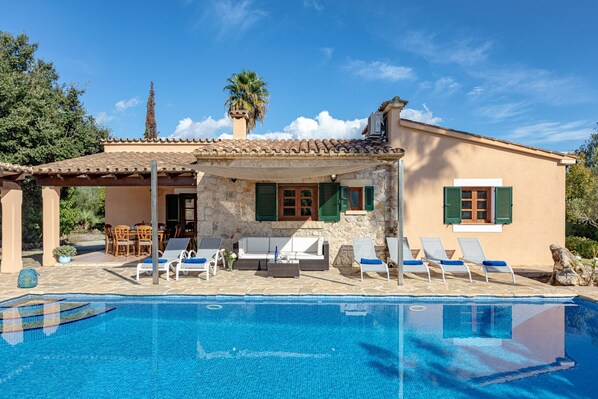 A poolside area with lounge chairs and a stone building with green shutters. - Majorca holiday rental - Villa Joana (Pollensa / Pollença)