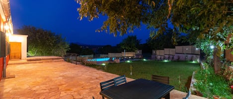 En udendørs terrasse med flisebelægning, et bord og stole, samt en swimmingpool i baggrunden.