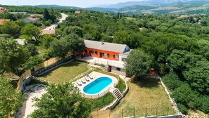 Una casa con piscina rodeada de jardín, ubicada en una zona montañosa con montañas al fondo.