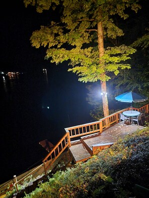 A wooden deck with a table and chairs, overlooking a body of water at night. - Waterfront Location. Panoramic Views. Swimming. Treehouse Tranquility. Private.  (Colton)