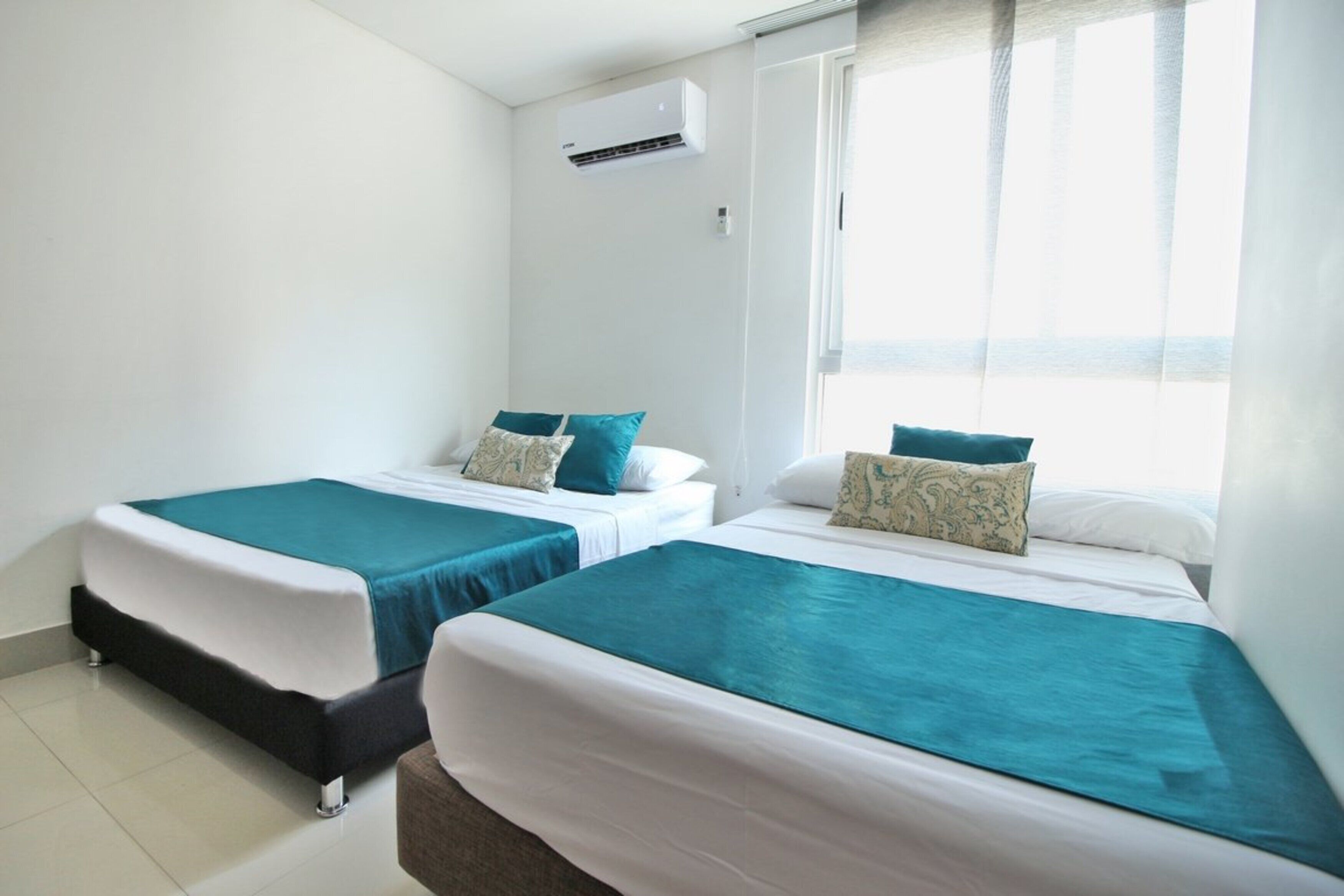 A hotel room with two beds, each with a teal bedspread and white pillows, a window with sheer curtains, and an air conditioning unit on the wall.