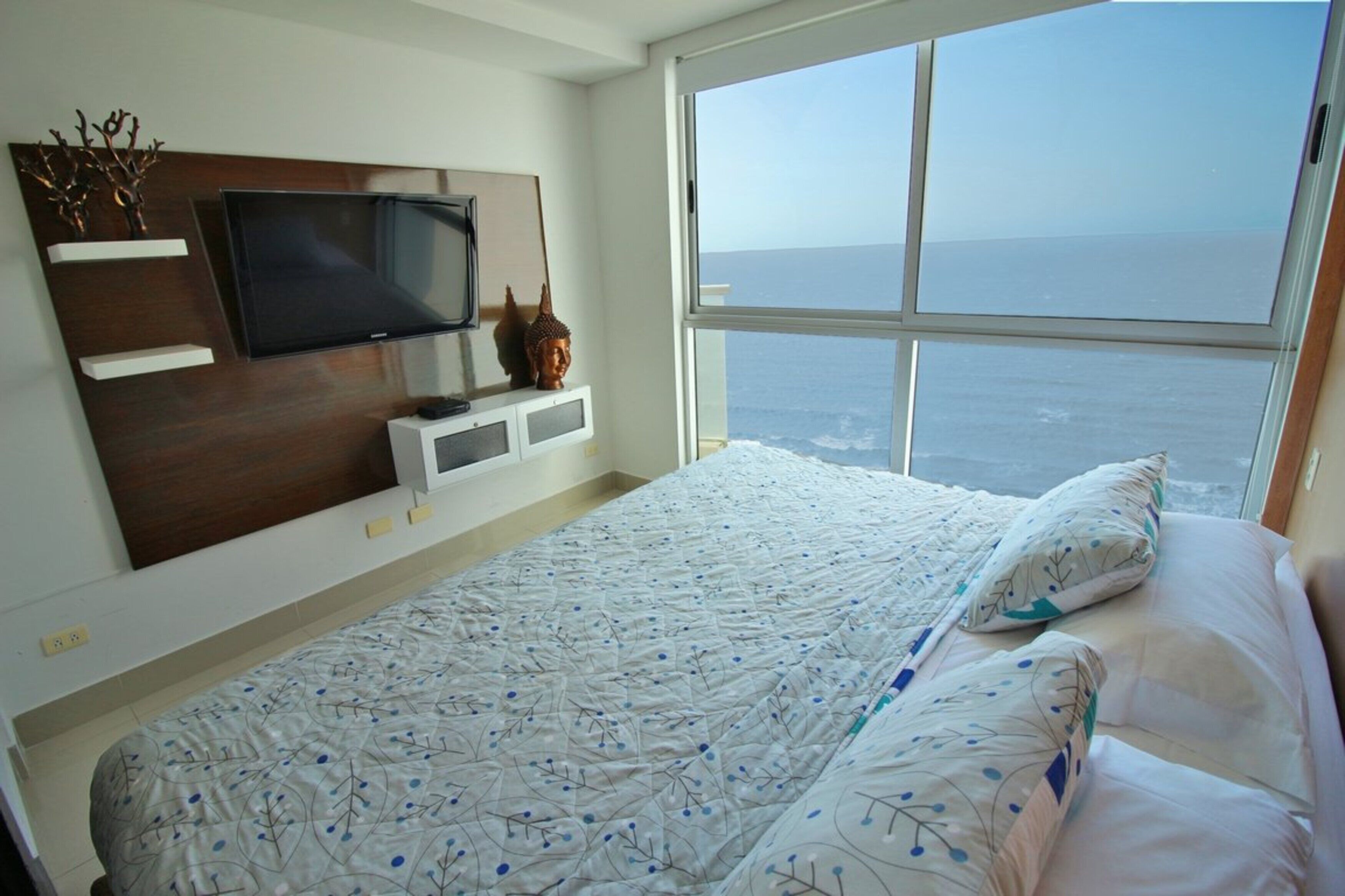 A bedroom with a large bed, a television on a wooden headboard, a window with a view of the sea, and a small statue on the nightstand.