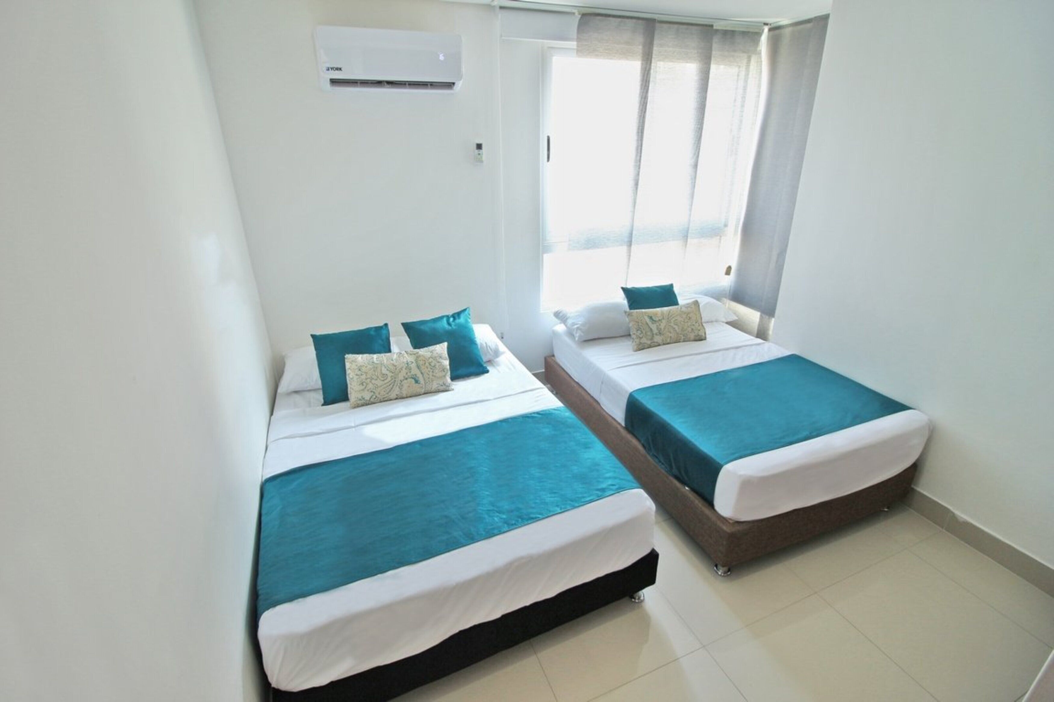 A hotel room with two beds, each with teal bedding and white pillows. There is a window with sheer curtains, and an air conditioning unit mounted on the wall.
