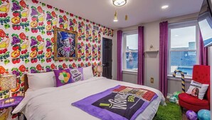Un dormitorio con un papel tapiz de frutas coloridas, una cama con una manta 'WONDERFUL BIRDS', un sillón rojo y una ventana con vistas a edificios.