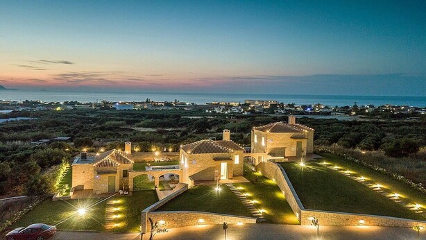 Ein Steinhaus mit beleuchteter Auffahrt und Blick auf das Meer im Dämmerlicht.