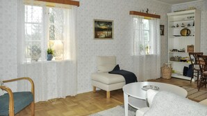 Un salon chaleureux avec un canapé, un fauteuil, une petite table et une bibliothèque. Il y a deux fenêtres avec des rideaux, un tableau au mur et une plante en pot sur le rebord de la fenêtre.