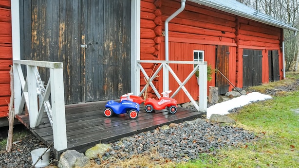 Une petite maison en rondins peinte en rouge, avec une véranda en bois et deux voitures jouets garées dessus.