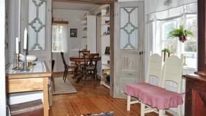 Une pièce avec un plancher en bois, une table à manger avec des chaises, une petite table recouverte d’une nappe à carreaux rouges et une fenêtre avec une plante.