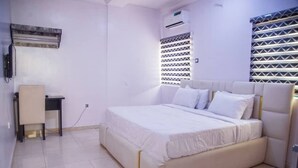 A bedroom with a bed, a desk, and a window with patterned blinds. - Blumerrit Hotel & Suites (Lagos)