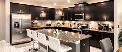 Una cocina moderna con armarios oscuros, electrodomésticos de acero inoxidable, una isla de granito con taburetes altos e iluminación colgante.