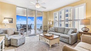 A spacious living room with a large sliding glass door leading to a balcony, a ceiling fan, and a view of the ocean. - Beach Colony East 10D - Indoor Pool - HASSLE-FREE CHECK OUT (Perdido Key)