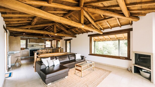 A spacious living area with a high wooden ceiling, a dark leather sofa, a wooden coffee table, and a large window providing natural light.