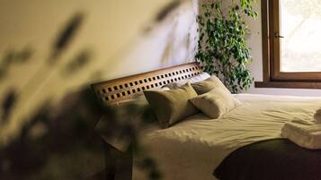 A neatly made bed with a wooden headboard, a potted plant beside it, and a window with natural light.