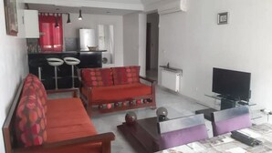 A living room with a sofa, a coffee table, and a television. There is a kitchen area in the background with a refrigerator and a stove. - House F. (Hammam Sousse)