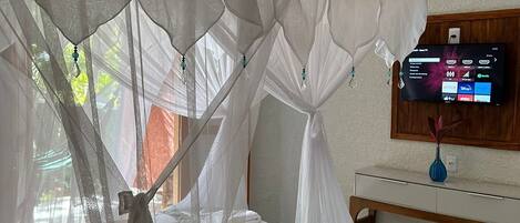 Un dormitorio con cama bajo un dosel blanco, una mesita de noche de madera con un florero y un televisor en la pared.