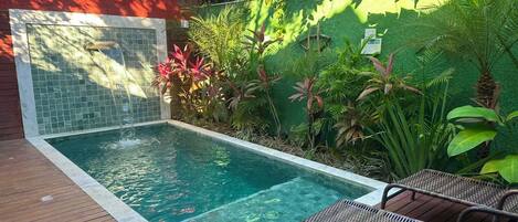 A swimming pool with a tiled wall, surrounded by lush greenery and a wooden deck with wicker chairs.