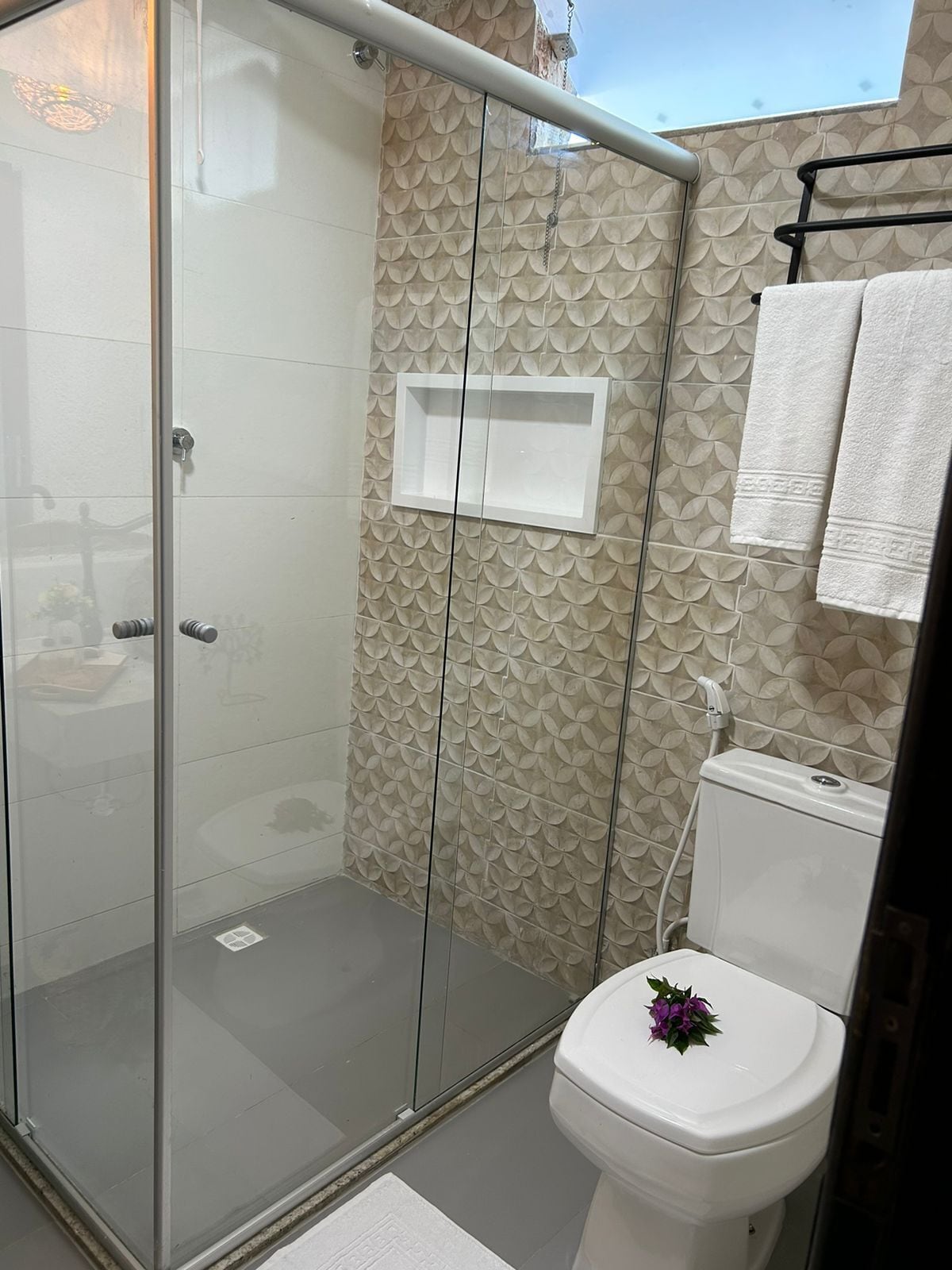 A modern bathroom with a glass shower enclosure, a toilet with a decorative flower on the lid, and a towel rack with white towels.